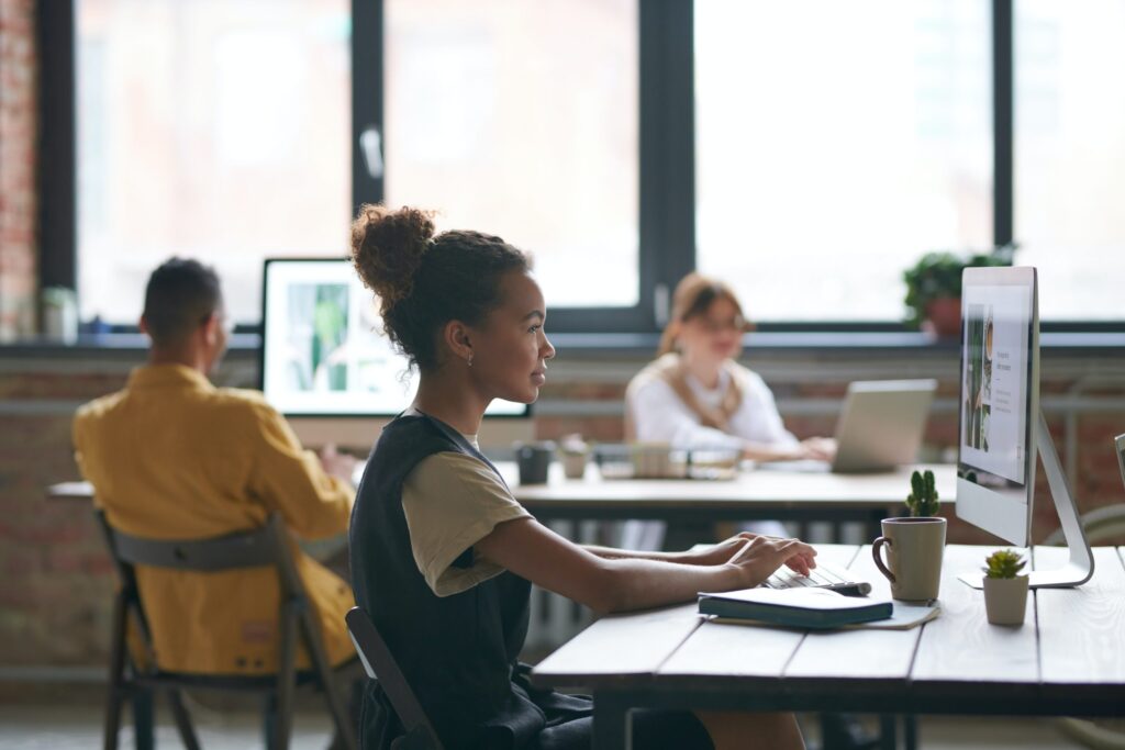 Woman working at a desktop computer in an open office with two colleagues in the background