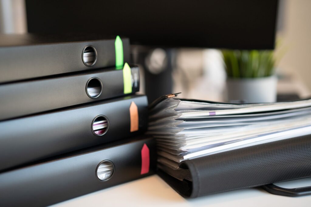 Stacks of black ring binders with colored tabs and a pile of documents on an office desk