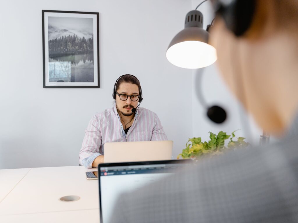 Man wearing a headset and glasses working on a laptop at a white desk in a bright office
