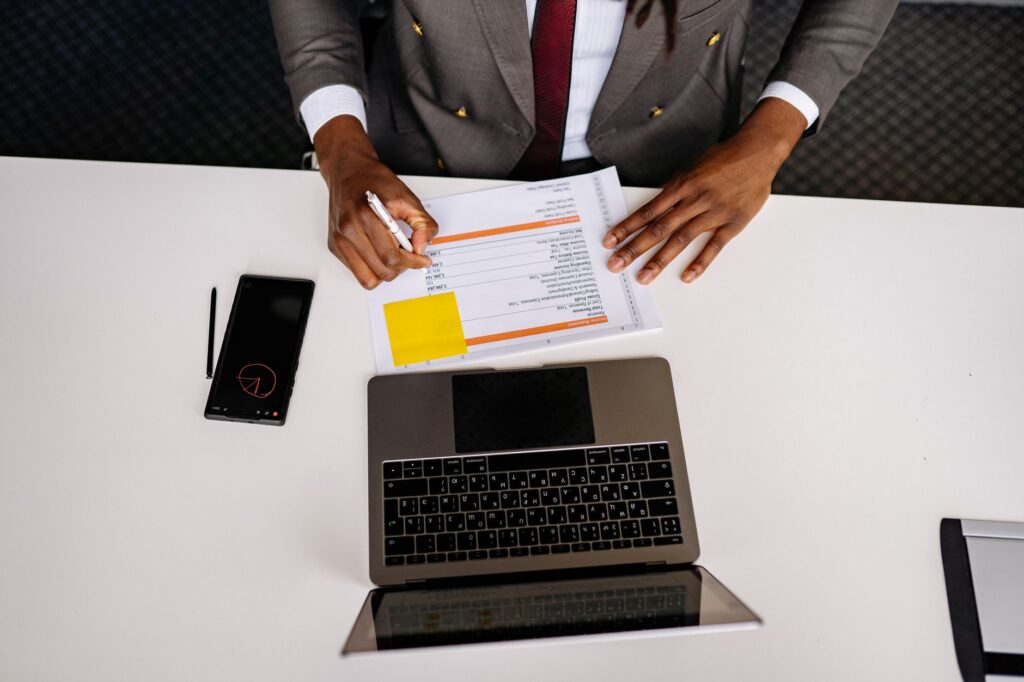 Overhead view of a person in a suit filling in a printed form at a white desk beside an open laptop and a phone