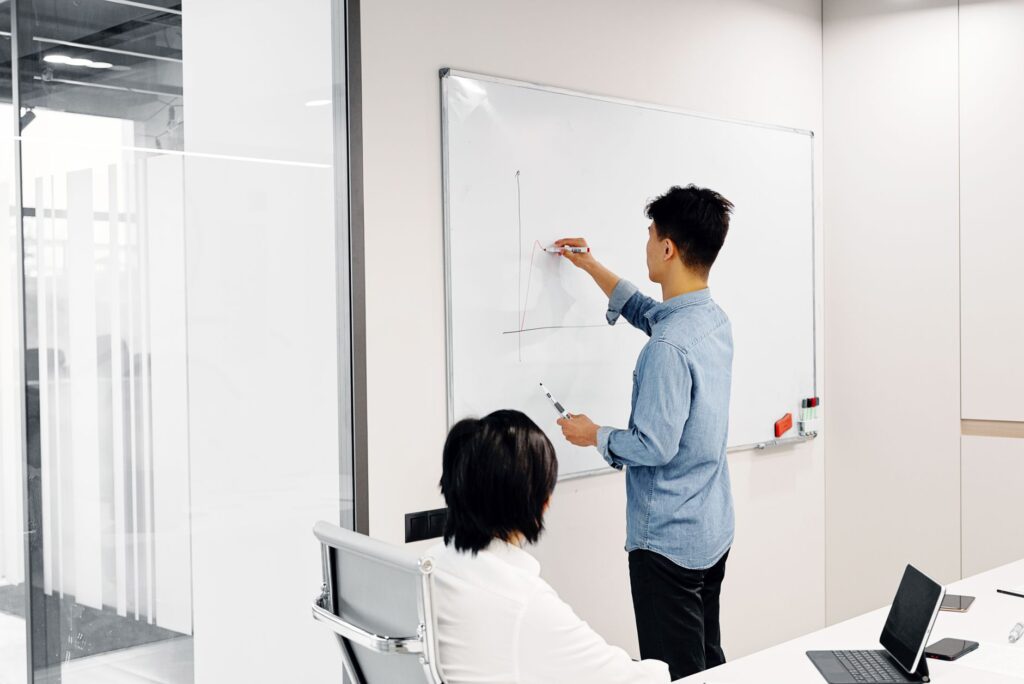 Man drawing a graph on a whiteboard while a seated colleague watches in a bright meeting room