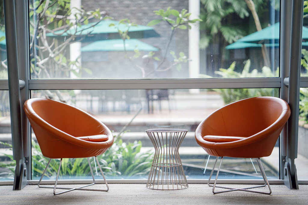 Two orange armchairs and a small table beside a glass wall overlooking a garden